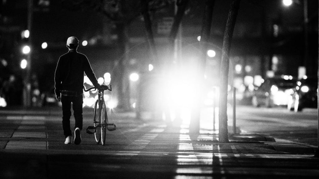 A bike at night silhouetted against headlights, which emphasizes how tricky visibility can be in Oregon and how bicyclists can protect themselves through the use of bike lights and bright or reflective clothing.