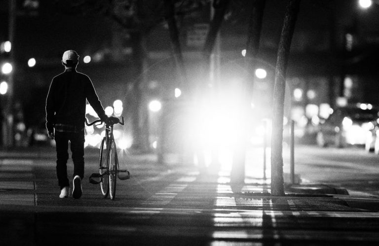 A bike at night silhouetted against headlights, which emphasizes how tricky visibility can be in Oregon and how bicyclists can protect themselves through the use of bike lights and bright or reflective clothing.