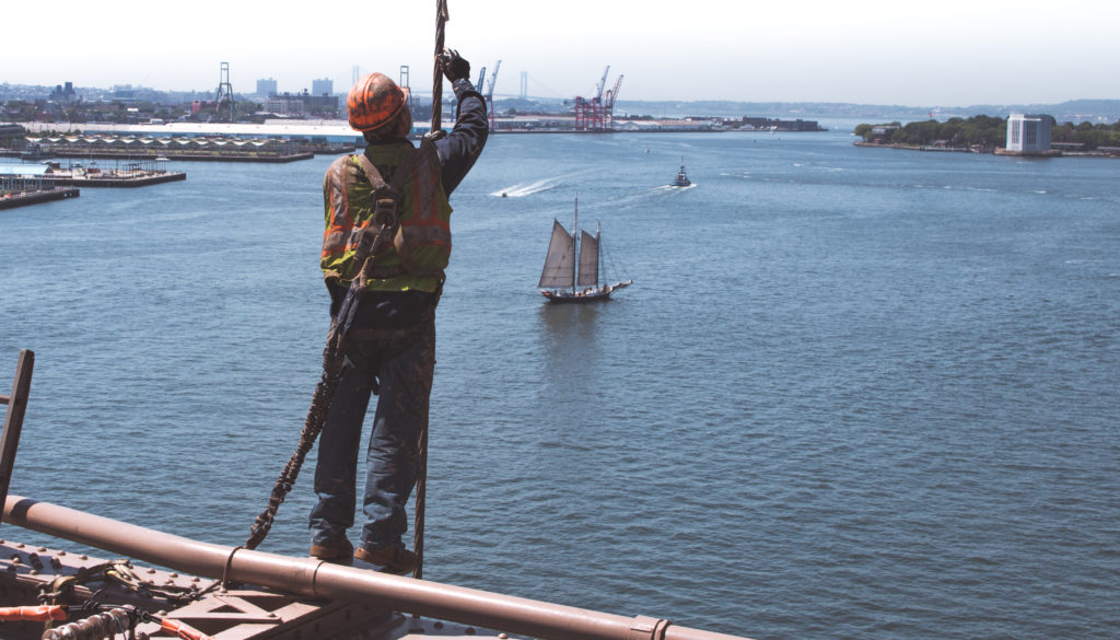 A construction worker on a bridge above water, wearing a hard hat and a safety harness as he works. Adequate construction site safety policies and practices are necessary to protect construction workers.