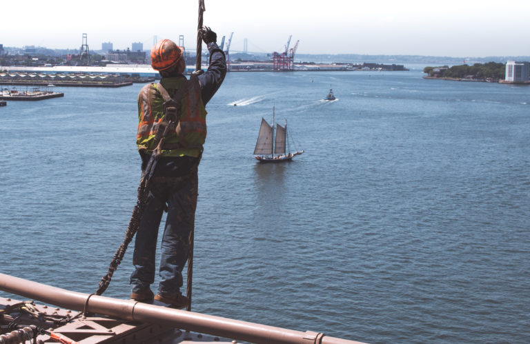 A construction worker on a bridge above water, wearing a hard hat and a safety harness as he works. Adequate construction site safety policies and practices are necessary to protect construction workers.