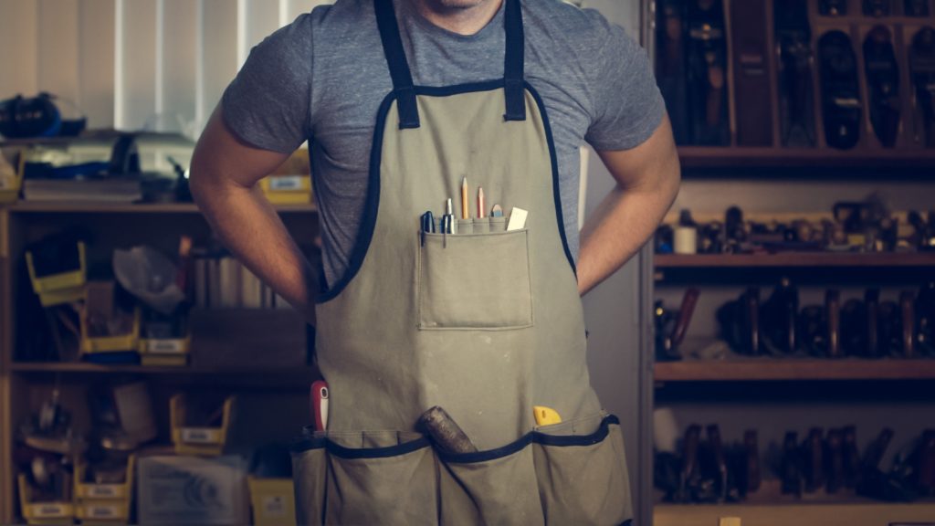 The torso of a white middle-aged male wearing an apron with woodworking tools.