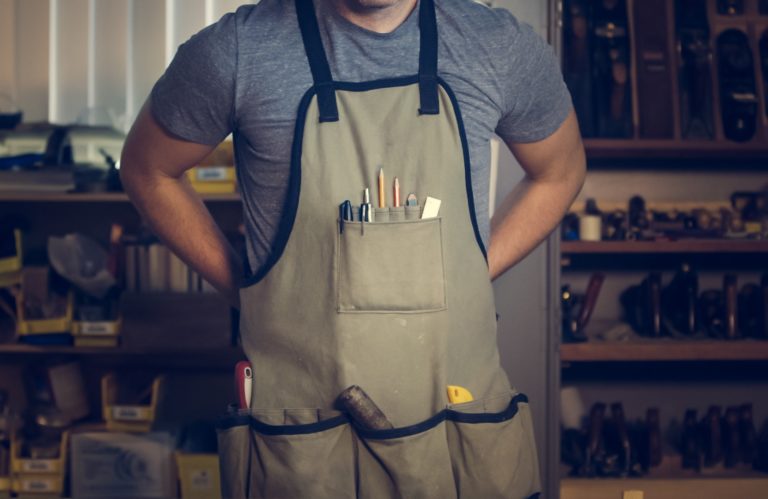 The torso of a white middle-aged male wearing an apron with woodworking tools.